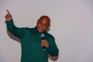 Speaker mid-speech with his index finger raised in one hand and a microphone in the other